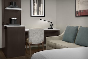 Une chambre d’hôtel avec un lit, un bureau doté d’une machine à café, une chaise, un canapé et un tableau représentant un paysage urbain accroché au mur.
