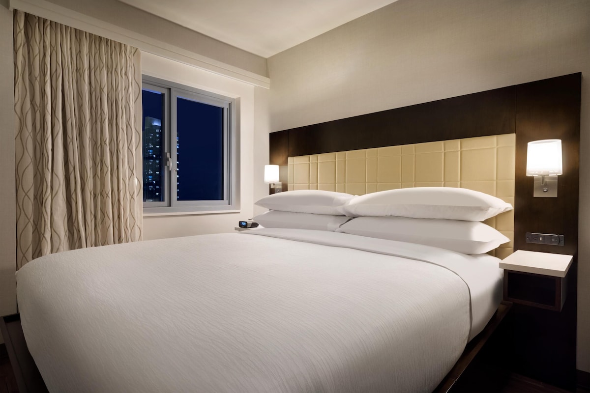 Een hotelkamer met een groot bed, een raam met gordijnen, een hoofdeinde met een lamp en een nachtkastje met een lampje.