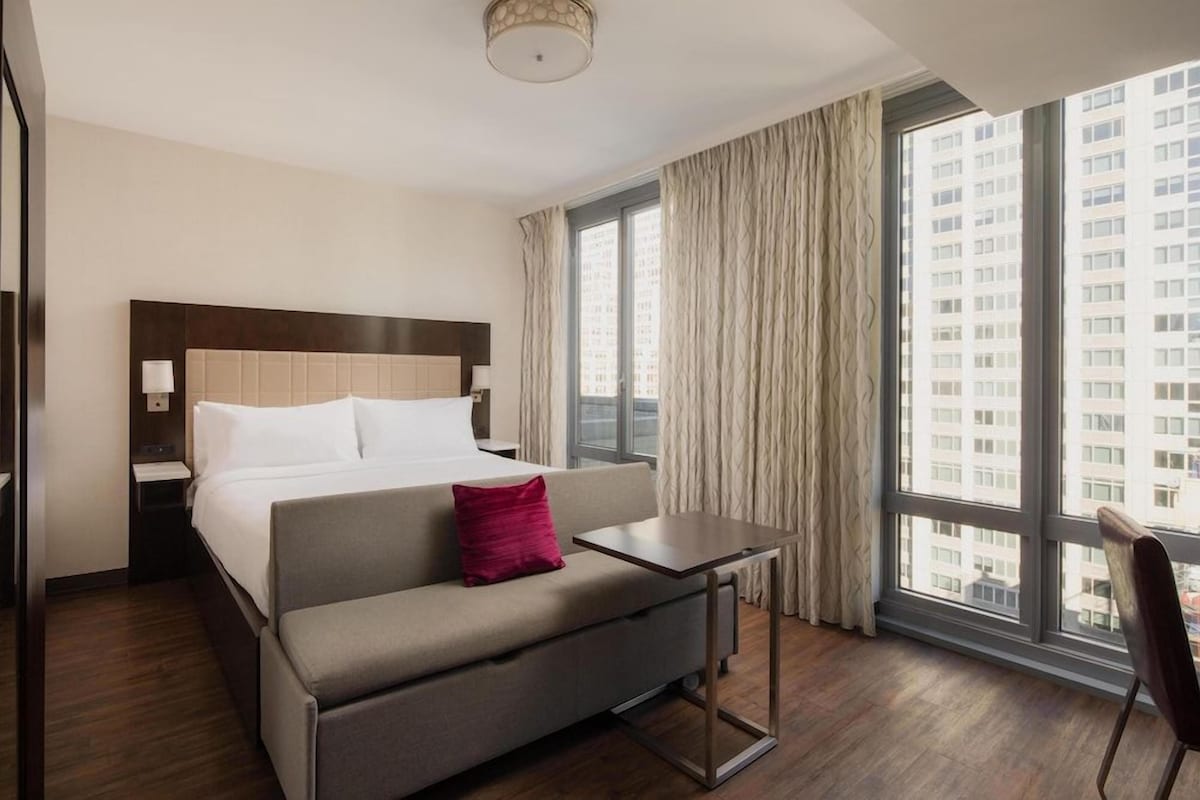 Quarto de hotel com cama, sofá, uma pequena mesa e janela com cortinas.