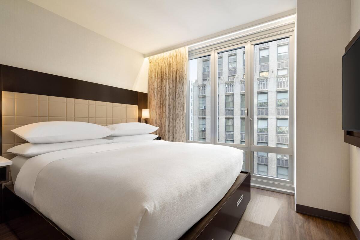 Une chambre d’hôtel moderne avec un grand lit, une fenêtre donnant sur des immeubles et un téléviseur à écran plat fixé au mur.