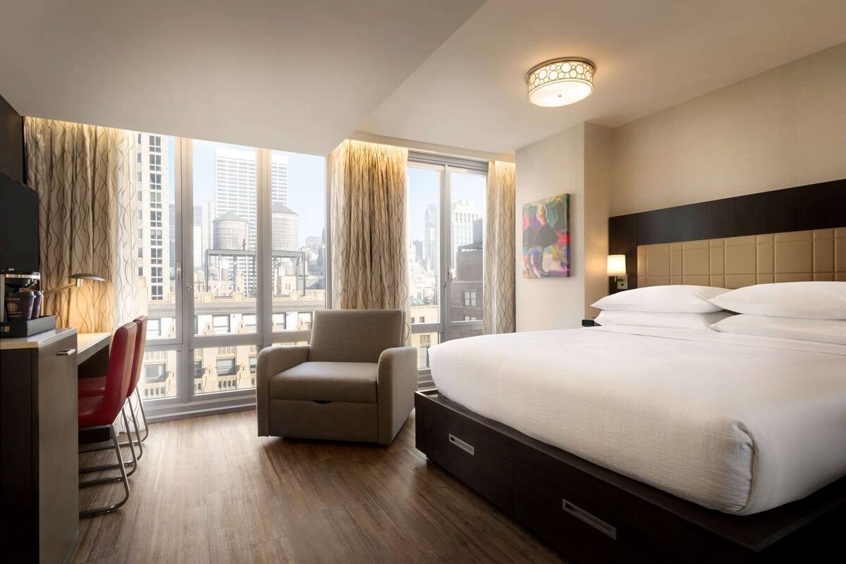 Une chambre d’hôtel équipée d’un lit, d’un bureau, d’une chaise et d’une grande fenêtre donnant sur la ville.