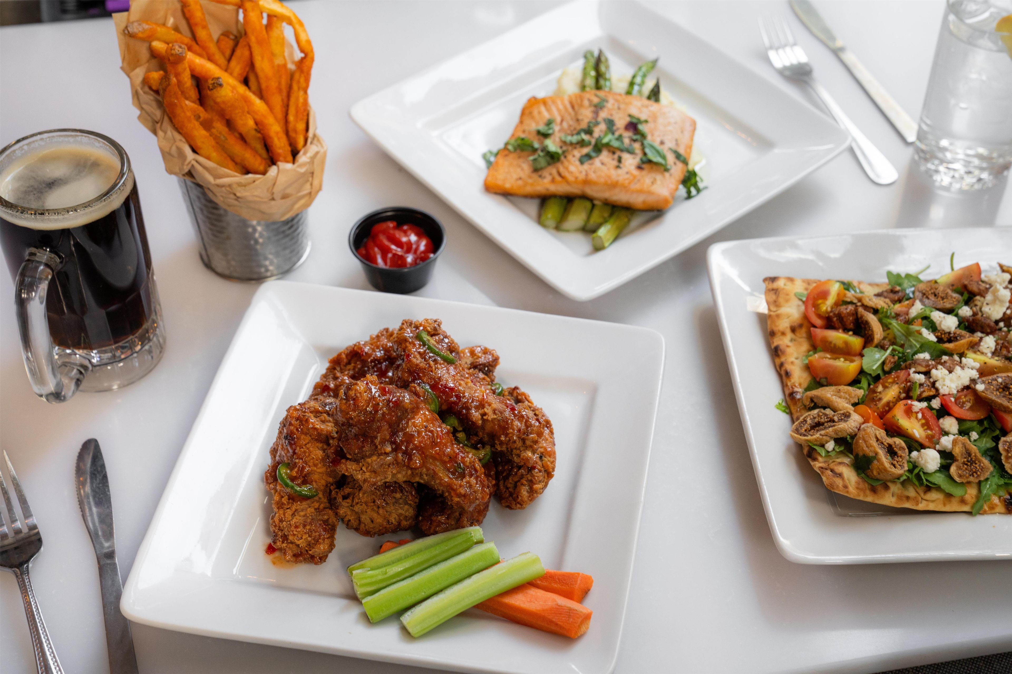 Une table sur laquelle on trouve divers plats, notamment une assiette d’ailes de poulet, une assiette de saumon, une pizza et un panier de frites.