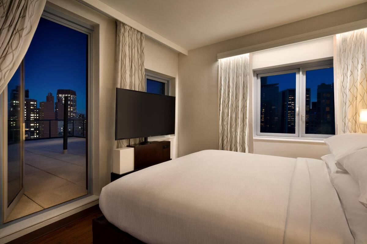 Une chambre d’hôtel avec un lit, une télévision, une fenêtre avec des rideaux et une vue sur le paysage urbain la nuit.