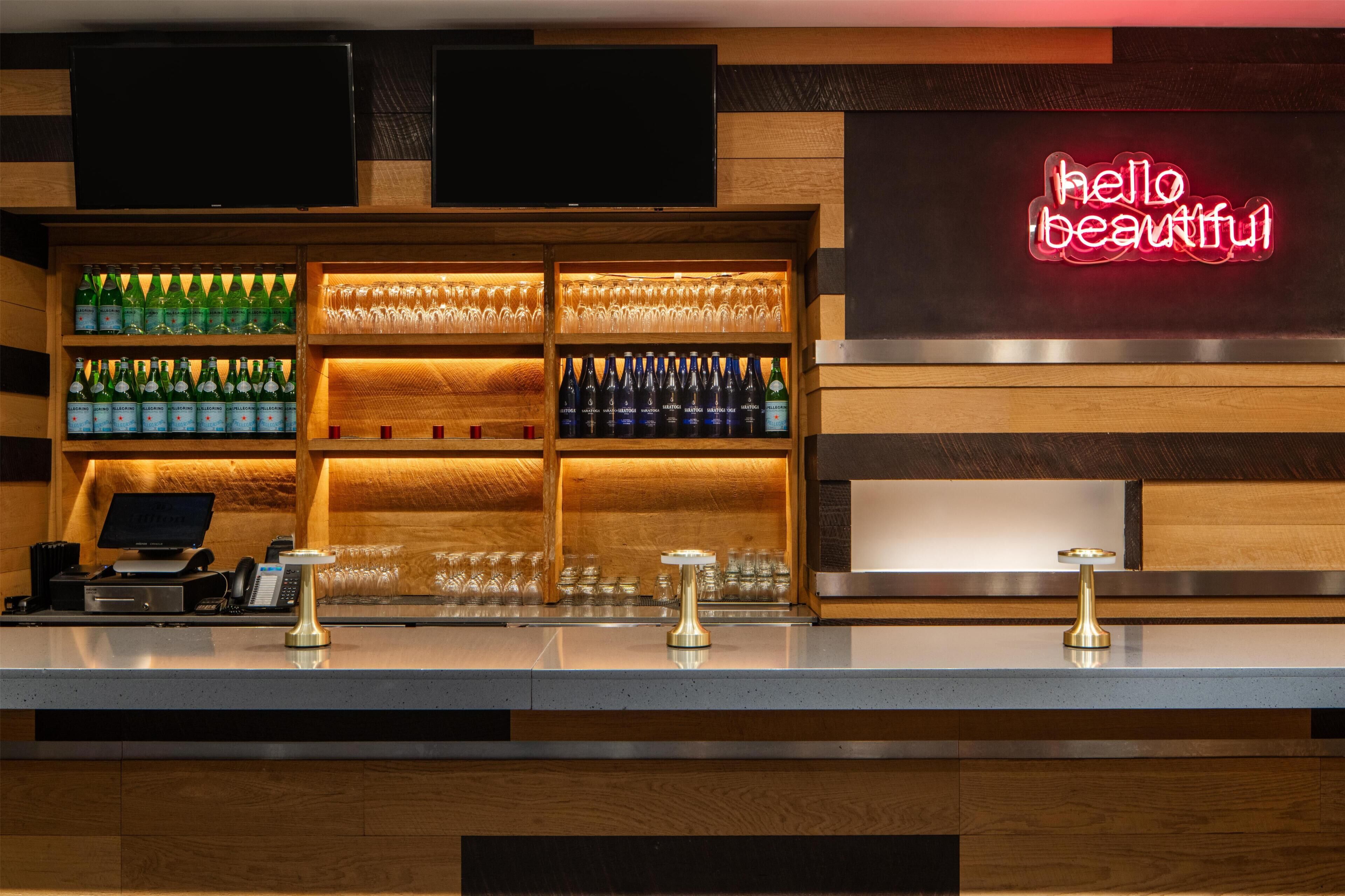 Zona de bar con estantes llenos de botellas y vasos, una caja registradora y un letrero de neón.
