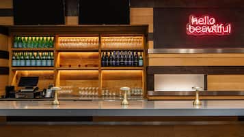 Zona de bar con estantes llenos de botellas y vasos, una caja registradora y un letrero de neón.