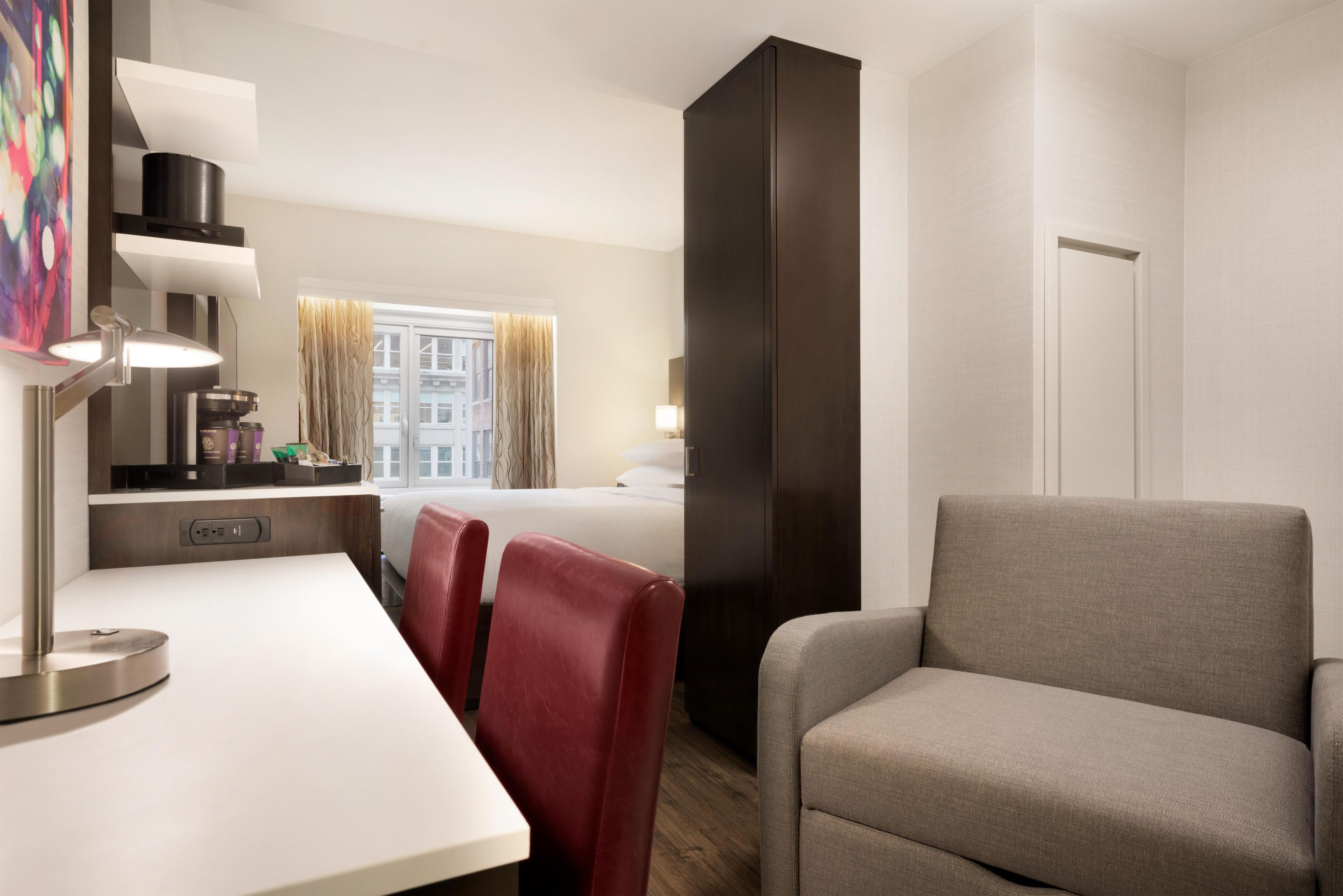 Una habitación de hotel con cama, escritorio con lámpara, silla, armario y ventana con cortinas.