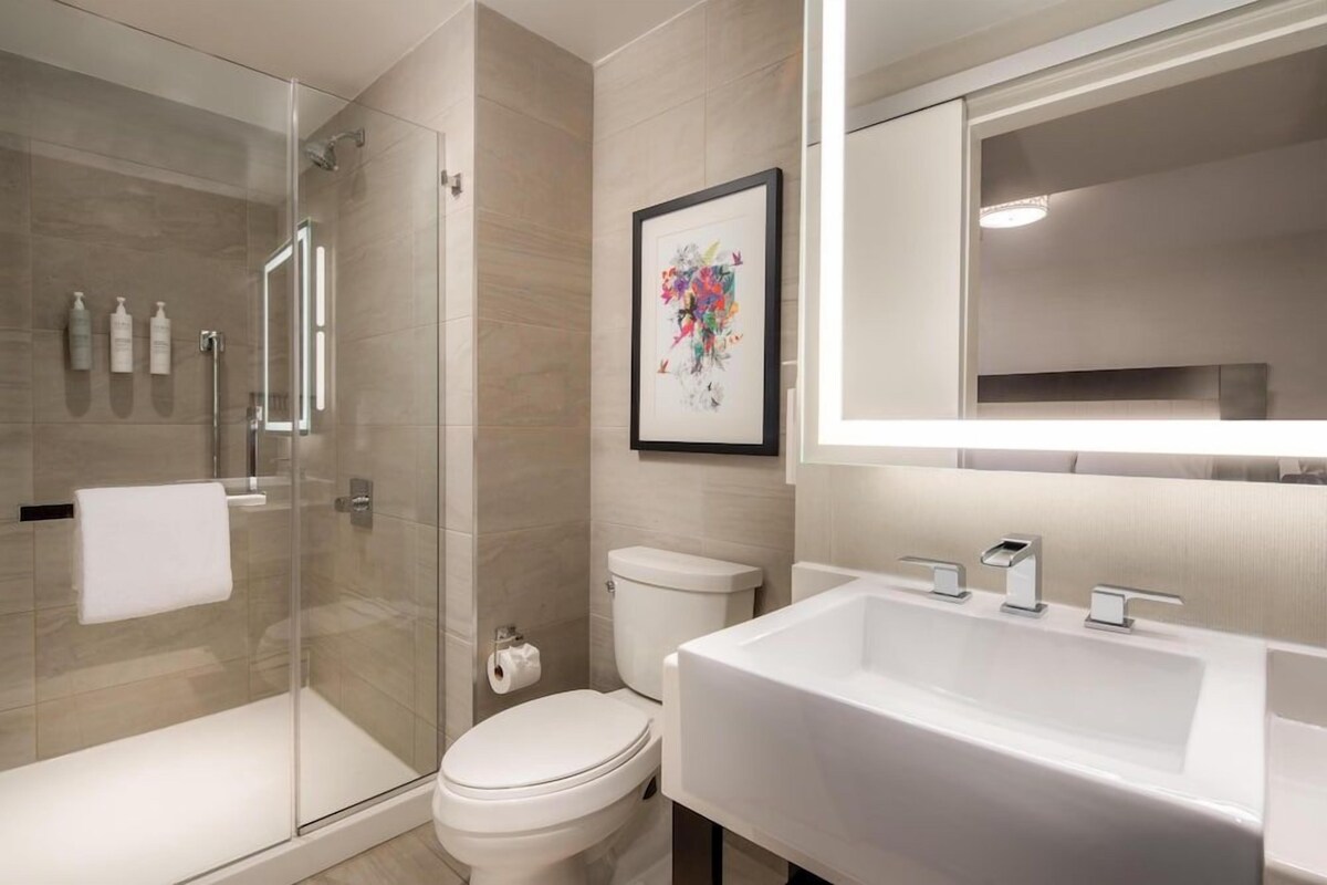 A modern bathroom with a glass shower enclosure, a white toilet, a large white sink, and a framed artwork on the wall.