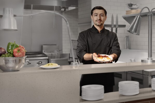 Un chef sostiene un plato de comida en una cocina moderna con electrodomésticos de acero inoxidable y un salpicadero de azulejos blancos.