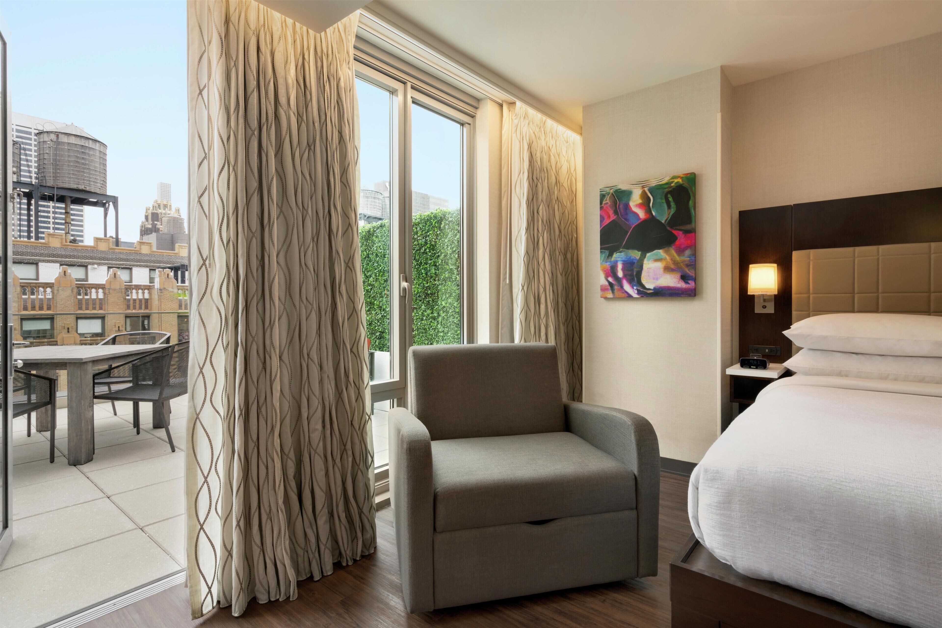 A hotel room with a bed, a chair, a table, and a painting on the wall.