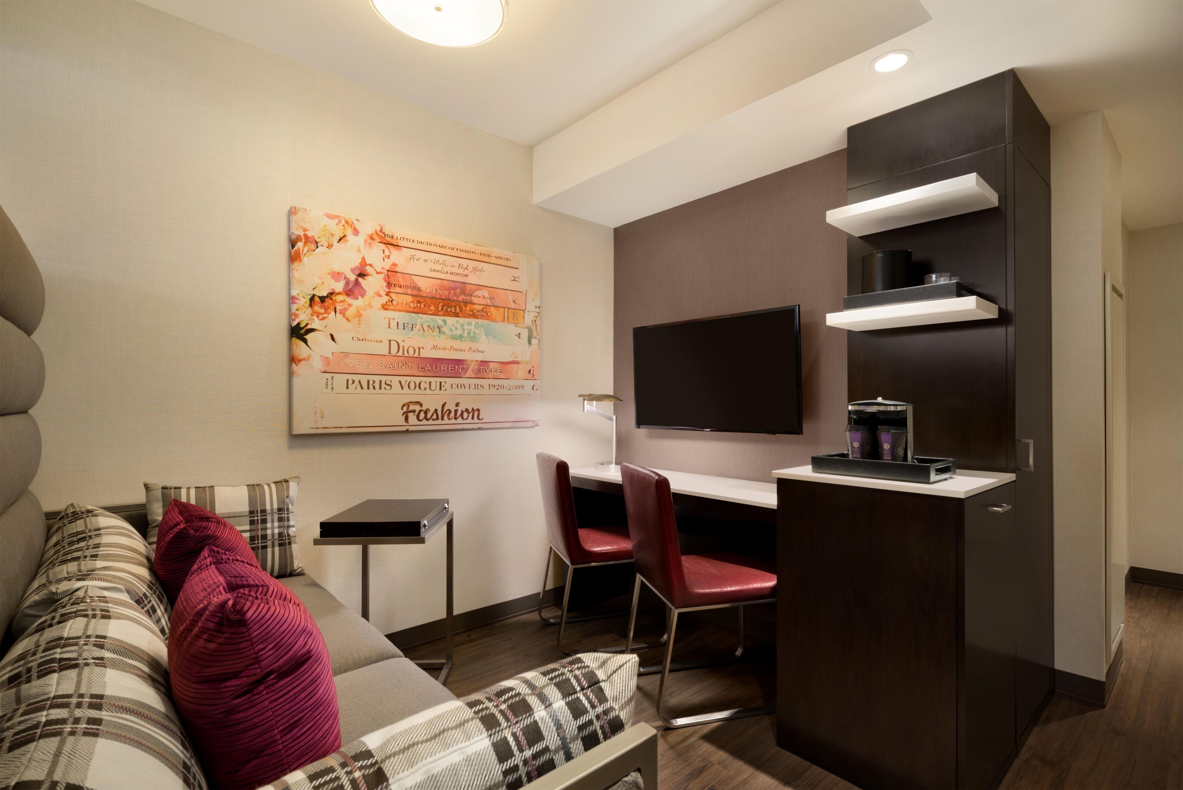A modern living room with a sofa, red and plaid cushions, a small table, a TV, and a built-in kitchen area with a microwave and coffee maker.