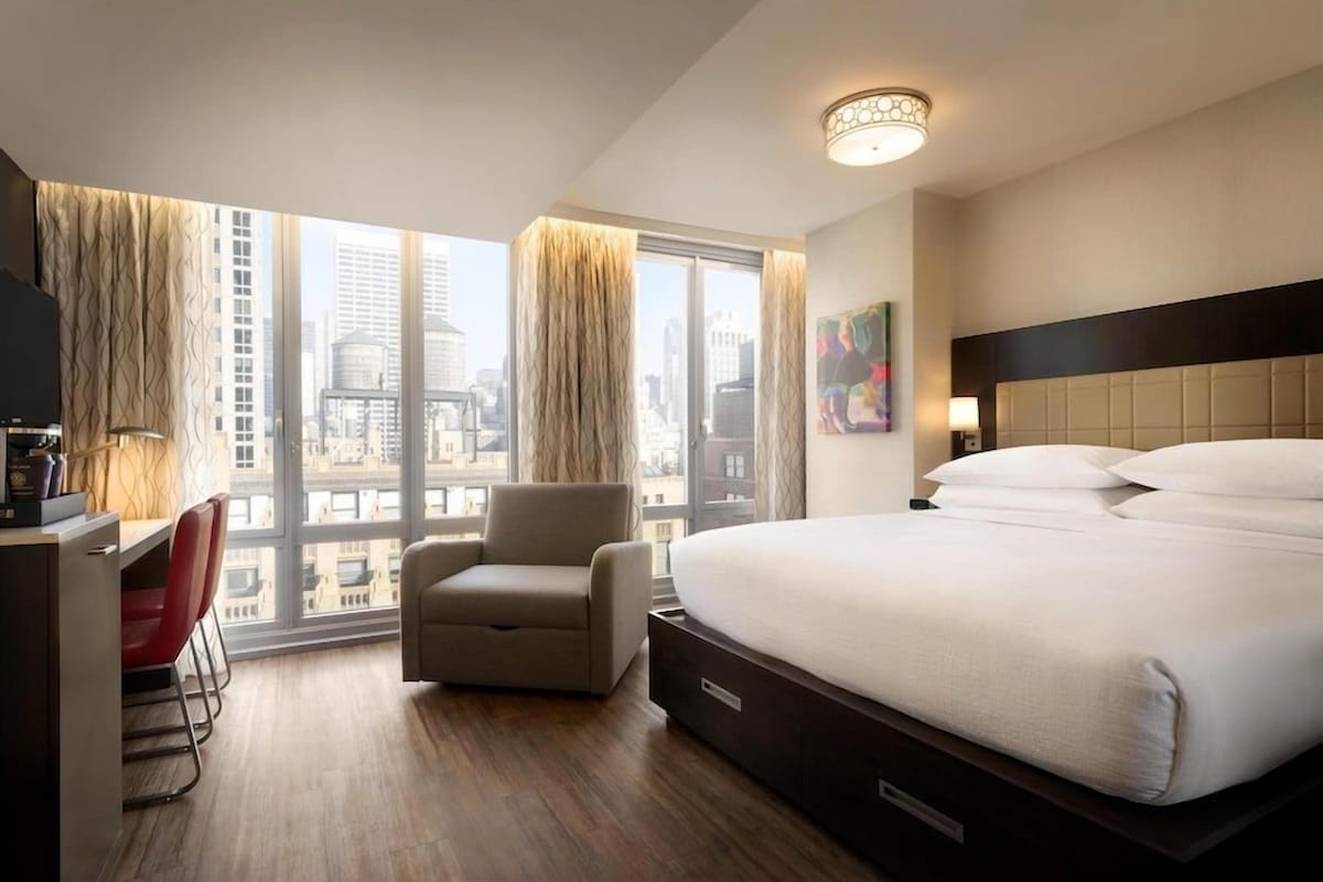 A hotel room with a large bed, a desk, a chair, and a city view.