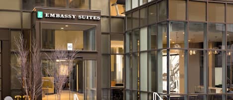 Un edificio moderno con un letrero que dice 'Embassy Suites'. Cuenta con amplios ventanales y se puede ver el interior con zonas de estar.
