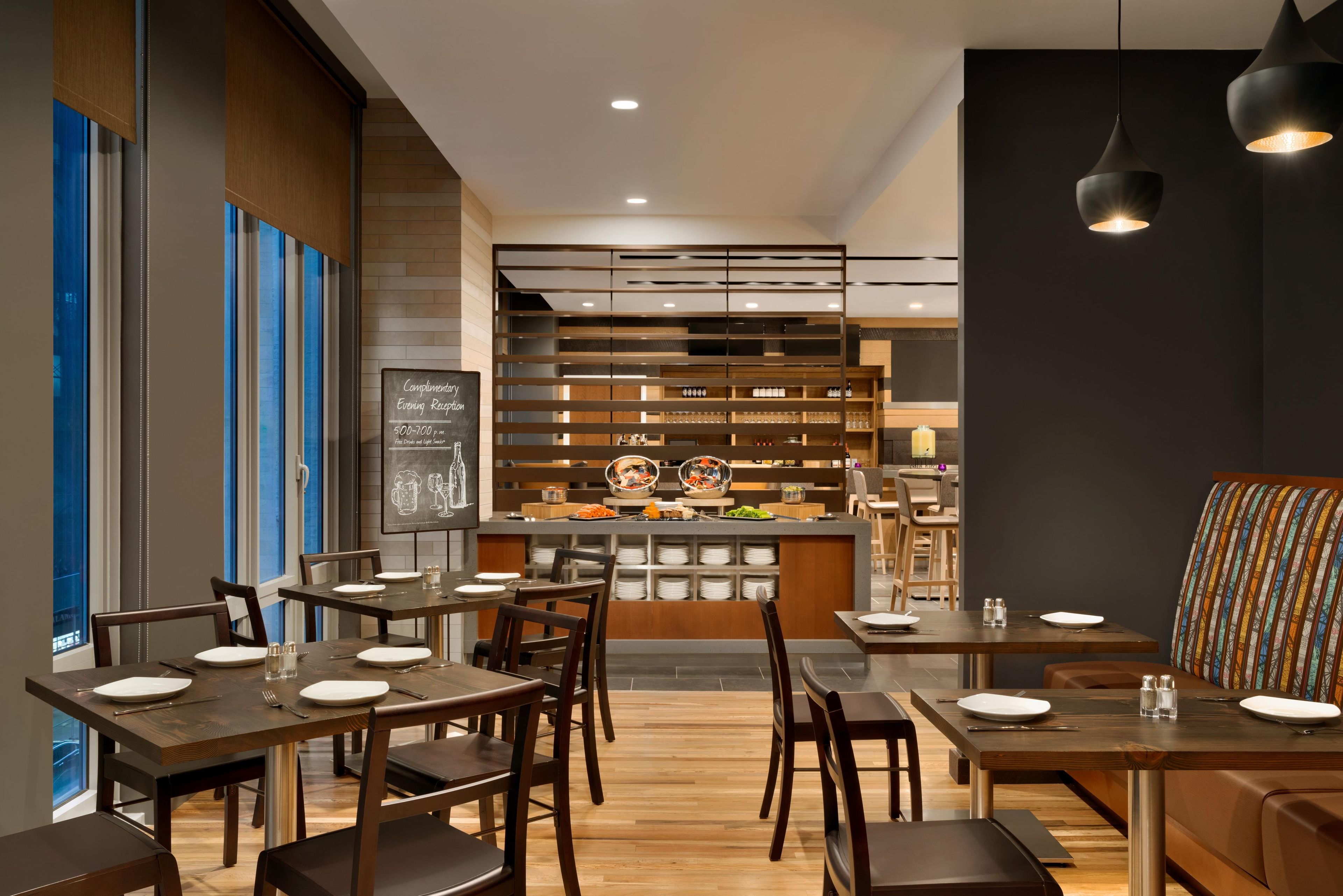 Un restaurante moderno con mesas y sillas de madera, una barra con menú, y un área de estar con un sofá colorido.
