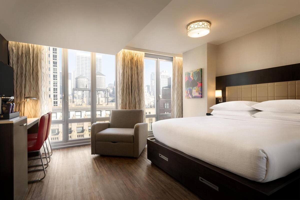Une chambre d’hôtel équipée d’un lit, d’un bureau, d’une chaise et d’une grande fenêtre donnant sur la ville.