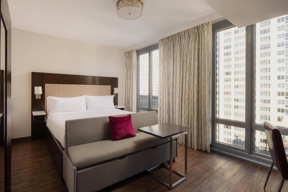 Une chambre d’hôtel avec un lit, un canapé, une petite table, une table de chevet et une fenêtre avec des rideaux.