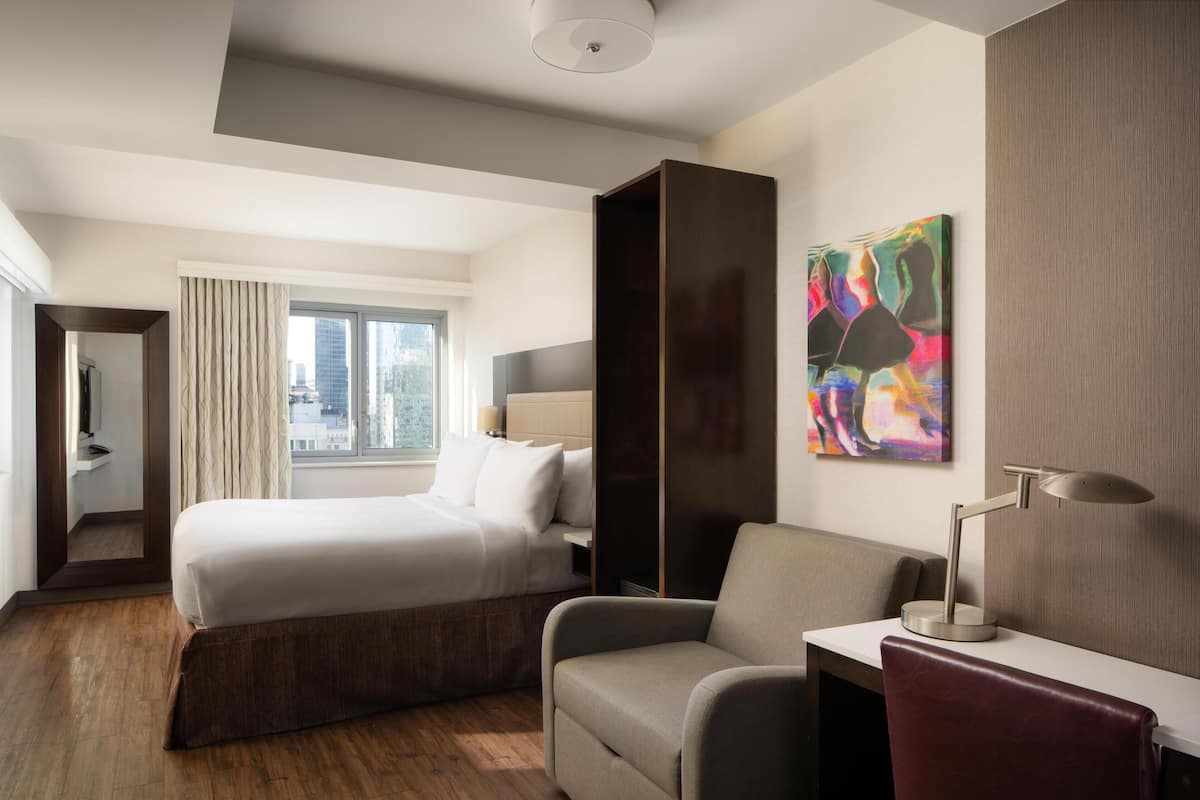 Une chambre d’hôtel avec un lit, un bureau, une chaise, une grande fenêtre et un tableau au mur.