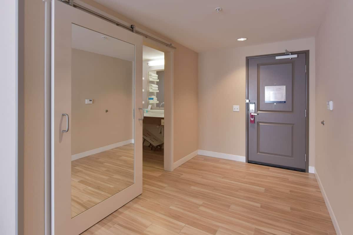 A hotel room with a wooden floor, a sliding door, and a visible bathroom with a sink and towel rack.