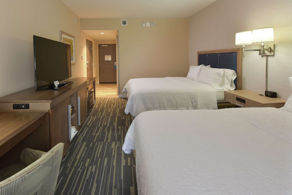 A hotel room with two beds, a desk with a flat-screen TV, a lamp, and a door leading to a hallway.