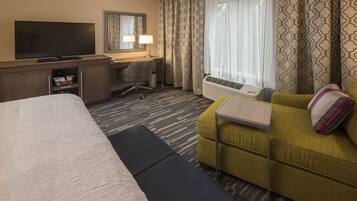 A hotel room with a bed, a desk with a chair, a television, a window with curtains, and a sofa.