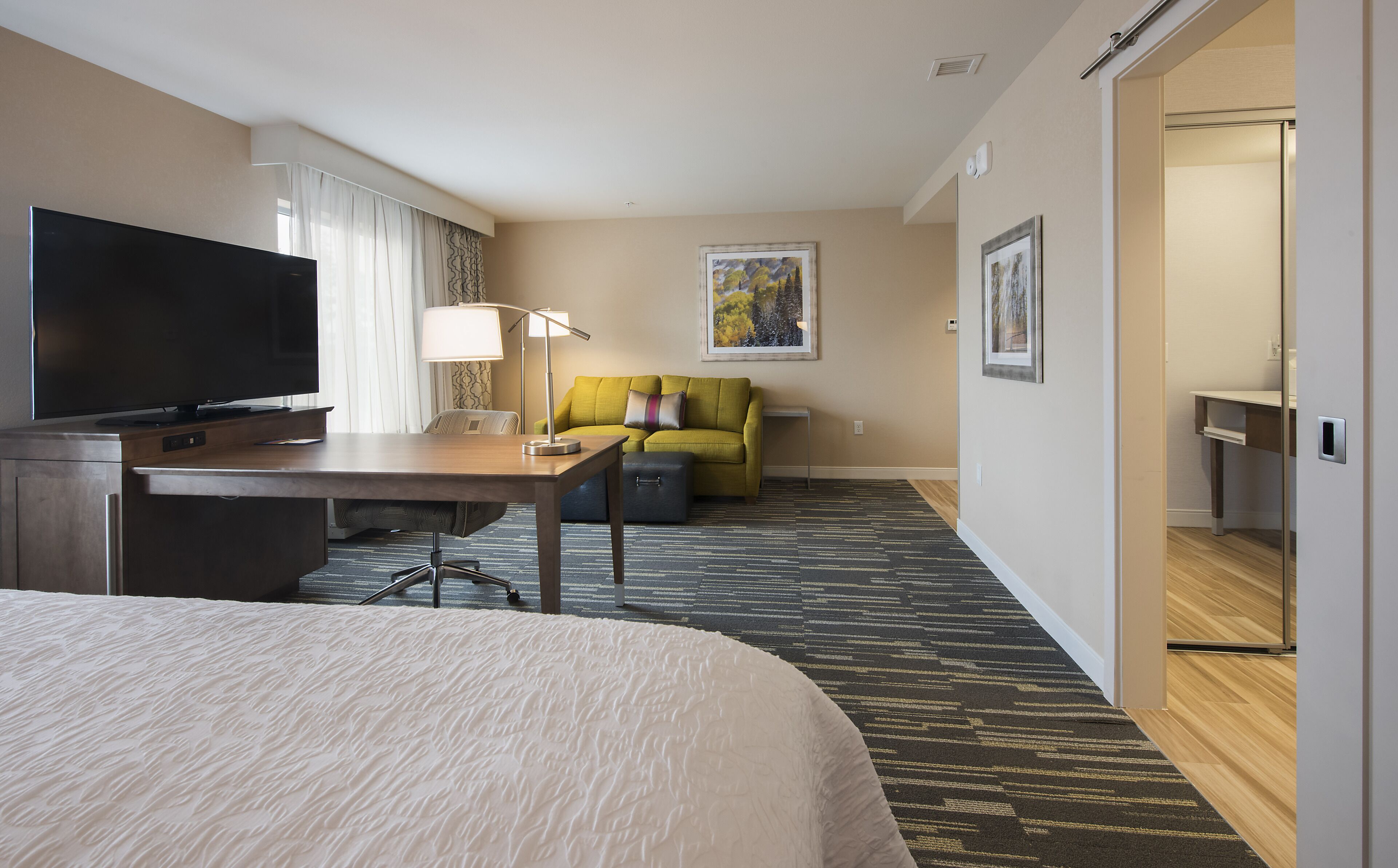 A hotel room with a bed, a desk with a flat-screen TV, a sofa, a lamp, and a painting on the wall.