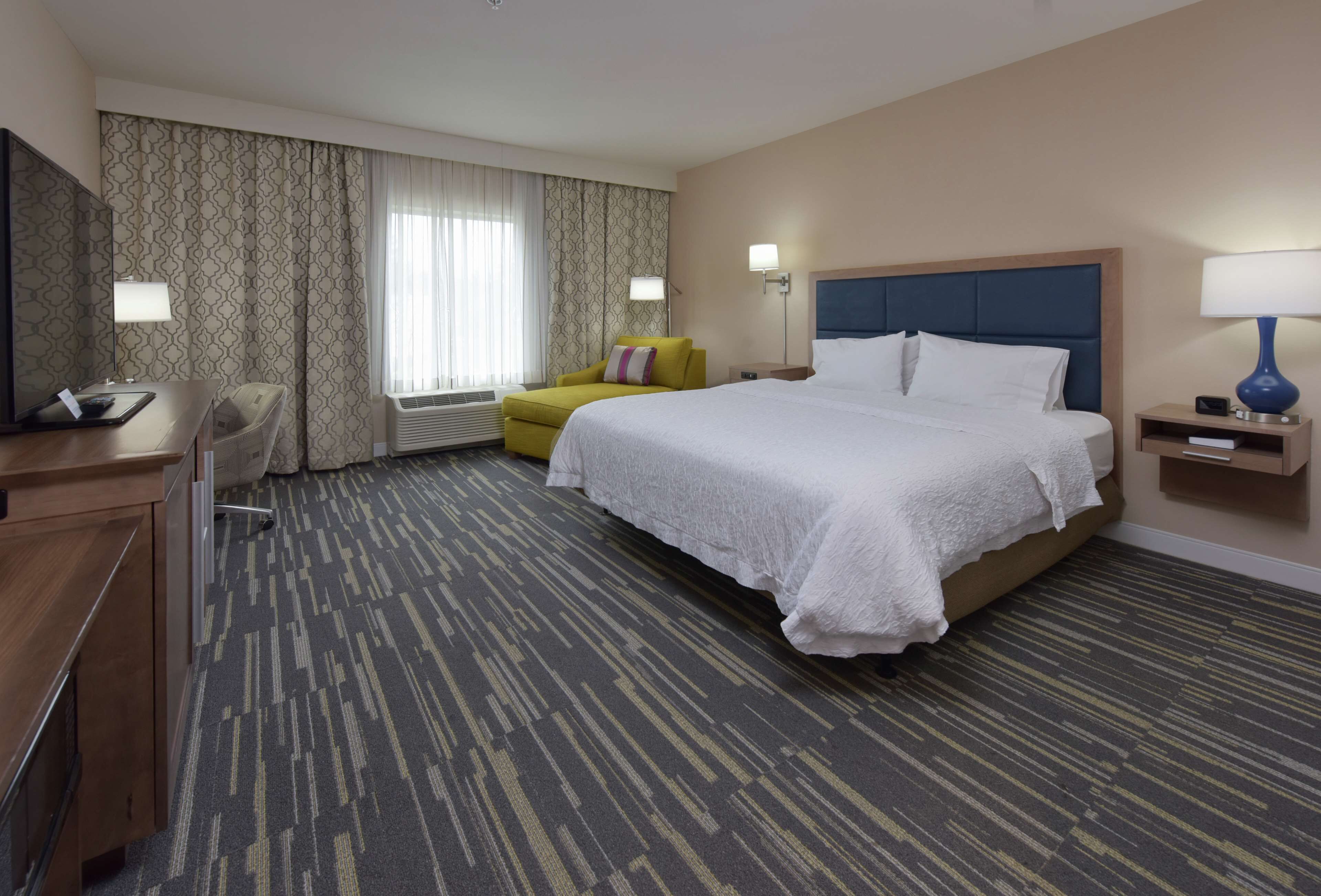 A hotel room with a large bed, a desk, a chair, a TV, and a window with curtains.