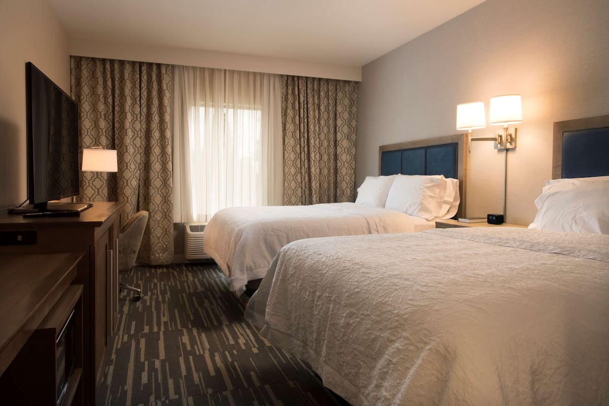 A hotel room with two beds, a desk with a computer, a flat-screen TV, a lamp, and a window with curtains.