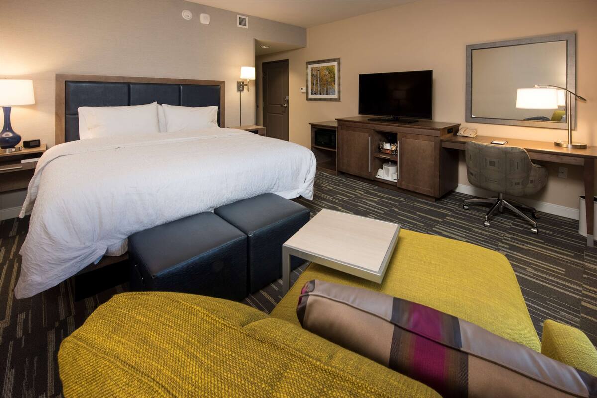 A hotel room with a large bed, a desk with a computer, a television, and a seating area with a sofa and ottoman.