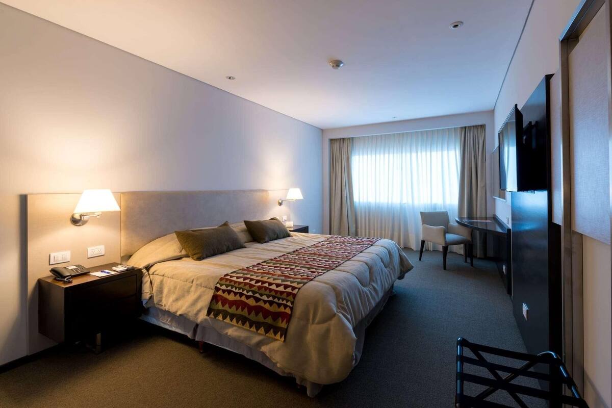 A hotel room with a bed, bedside lamps, a desk, a chair, and a TV.