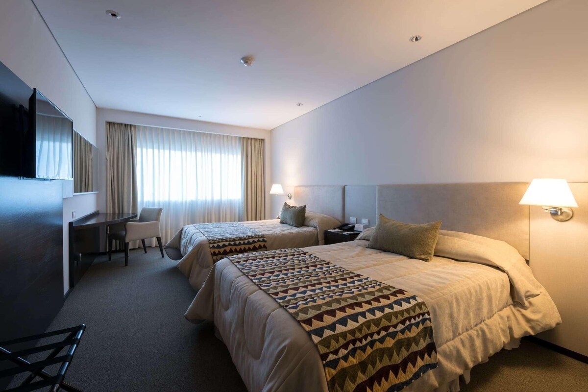 A hotel room with two beds, a desk, a chair, a TV, and a lamp.