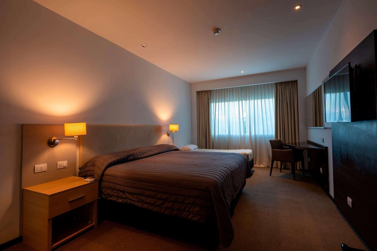 A hotel room with a bed, bedside lamps, a desk, a chair, and a television.
