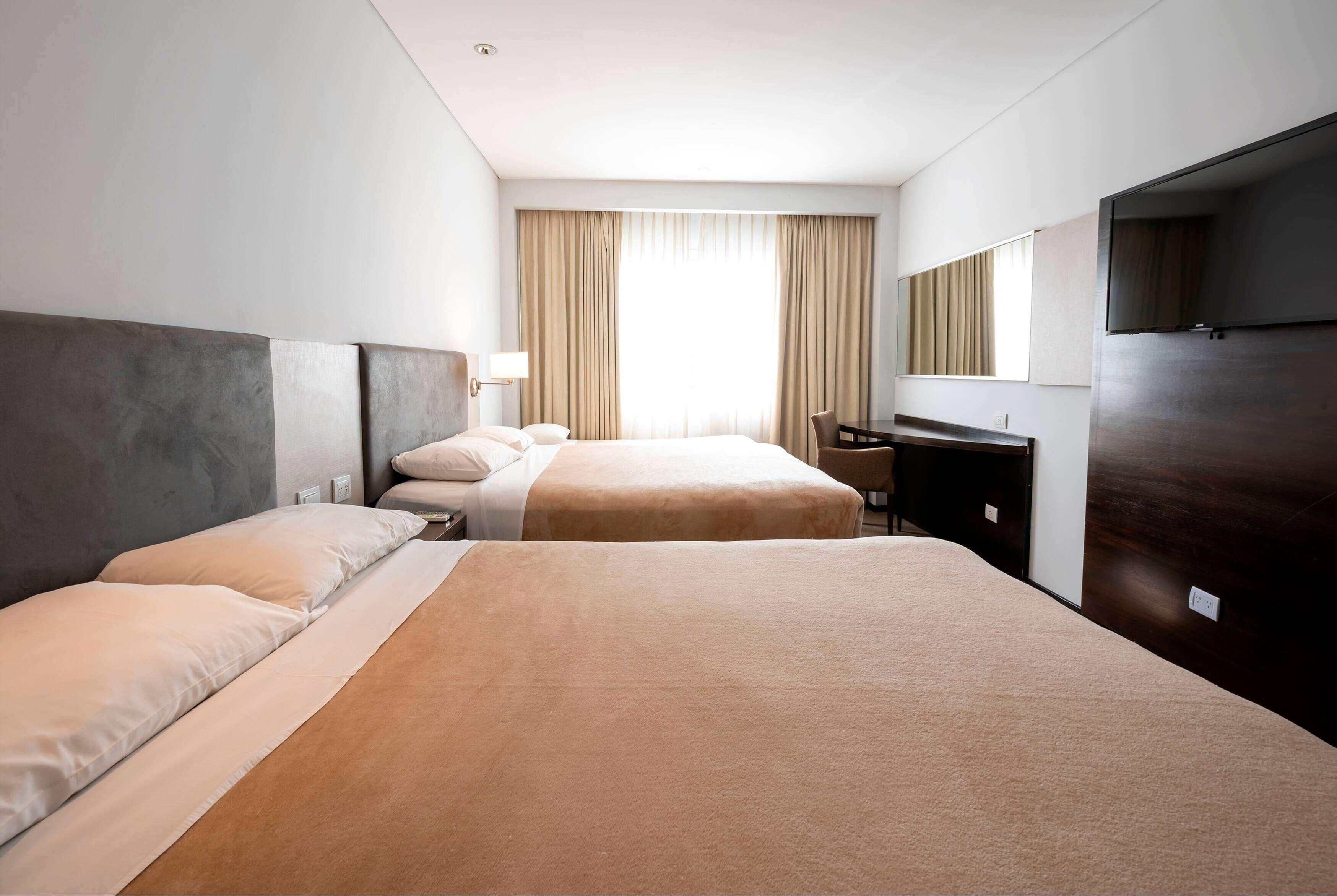 A hotel room with two beds, a desk, a chair, a TV, and a large window with curtains.
