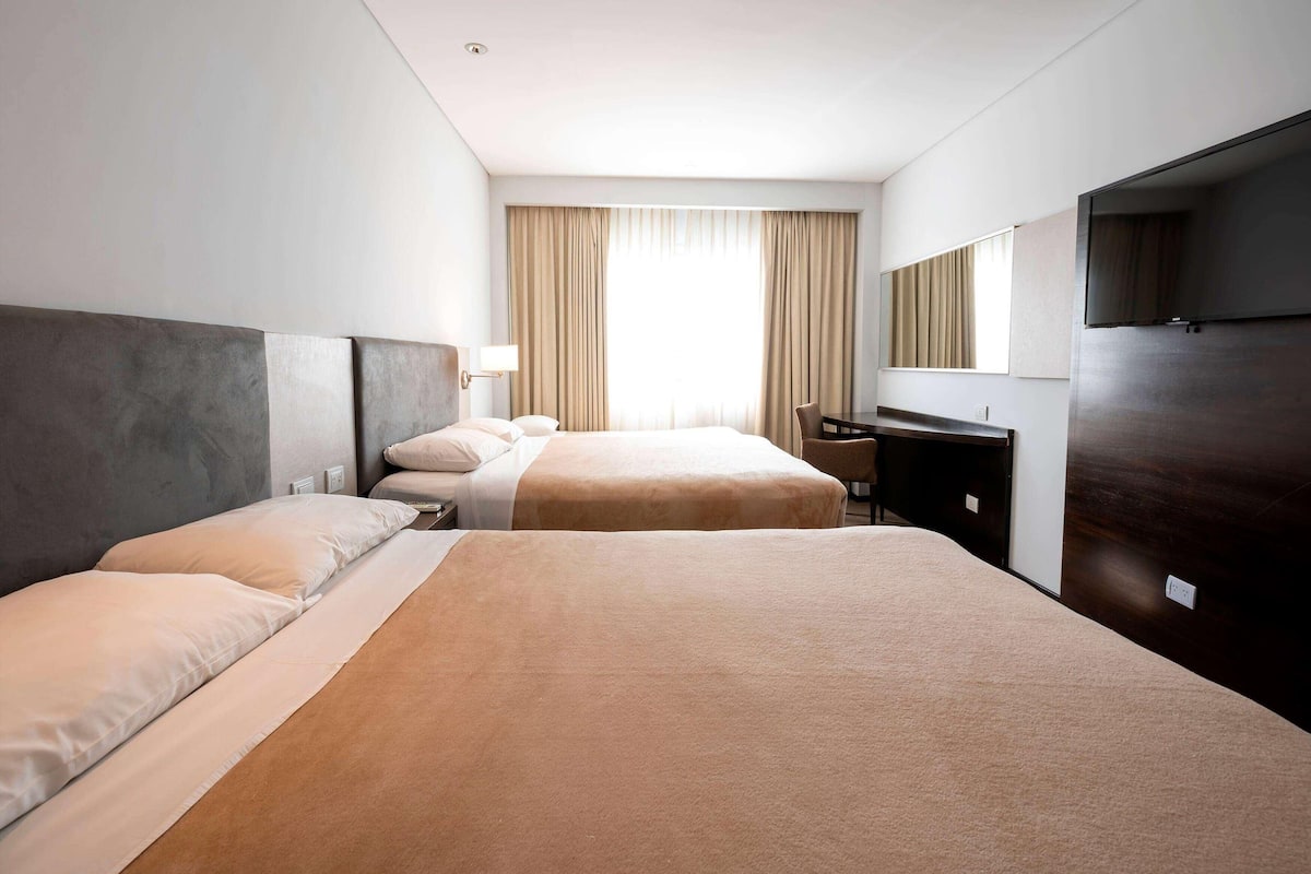 A hotel room with two beds, a desk, a chair, a TV, and a large window with curtains.
