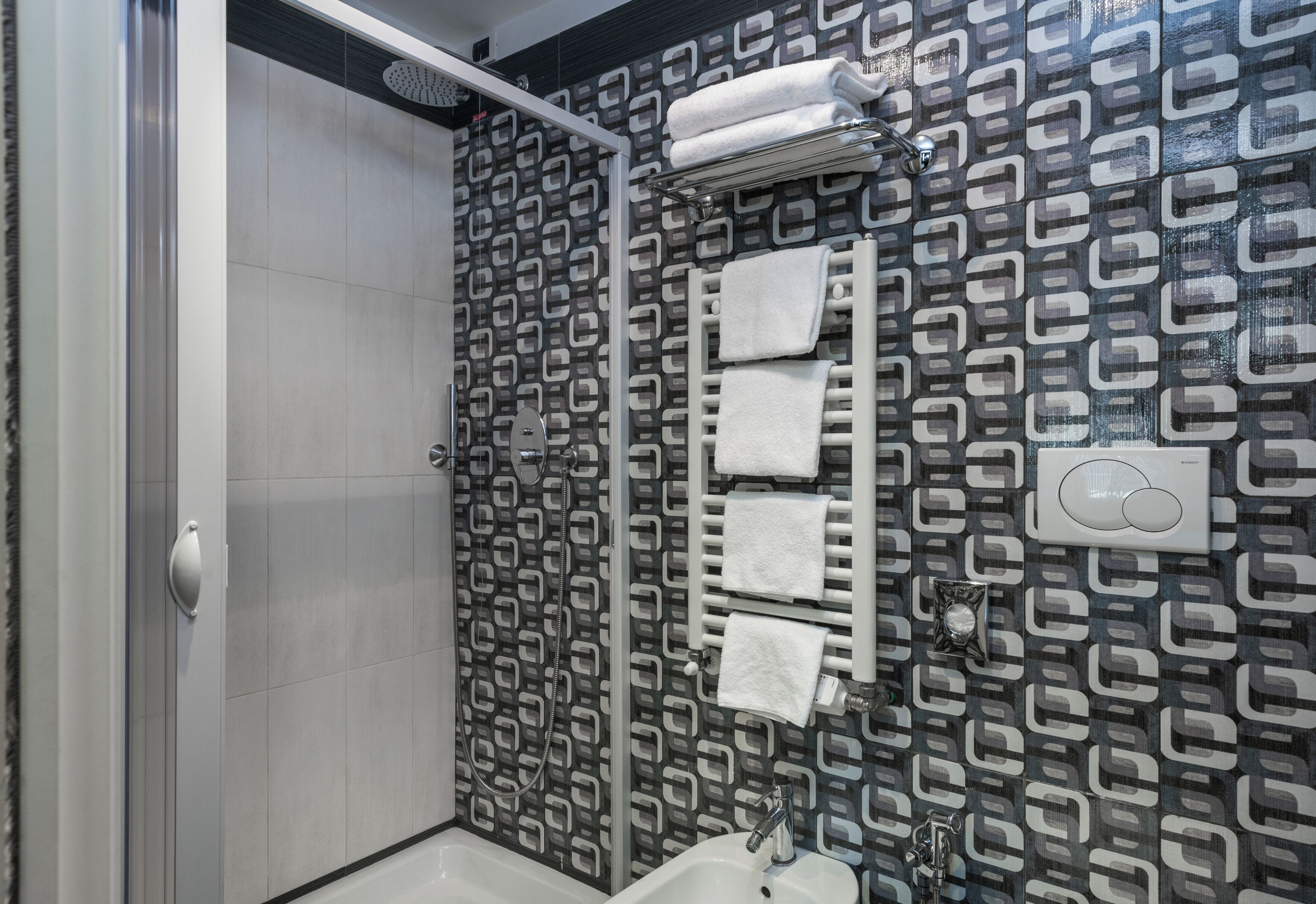 A modern bathroom with a patterned wall, a glass shower enclosure, a white sink, and a towel rack with multiple towels.