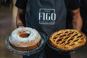 Egy személy, aki két desszertet – egy bundt-tortát és egy pitét – tart a kezében, és sötét színű kötényt visel, amelyen a „Ristorante FIGO” felirat olvasható.