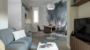 Ein modernes Wohnzimmer mit grauem Sofa, weißem Couchtisch und Flachbildfernseher. Es gibt eine Essecke mit Tisch und Stühlen sowie eine Bücherwand mit Büchern.