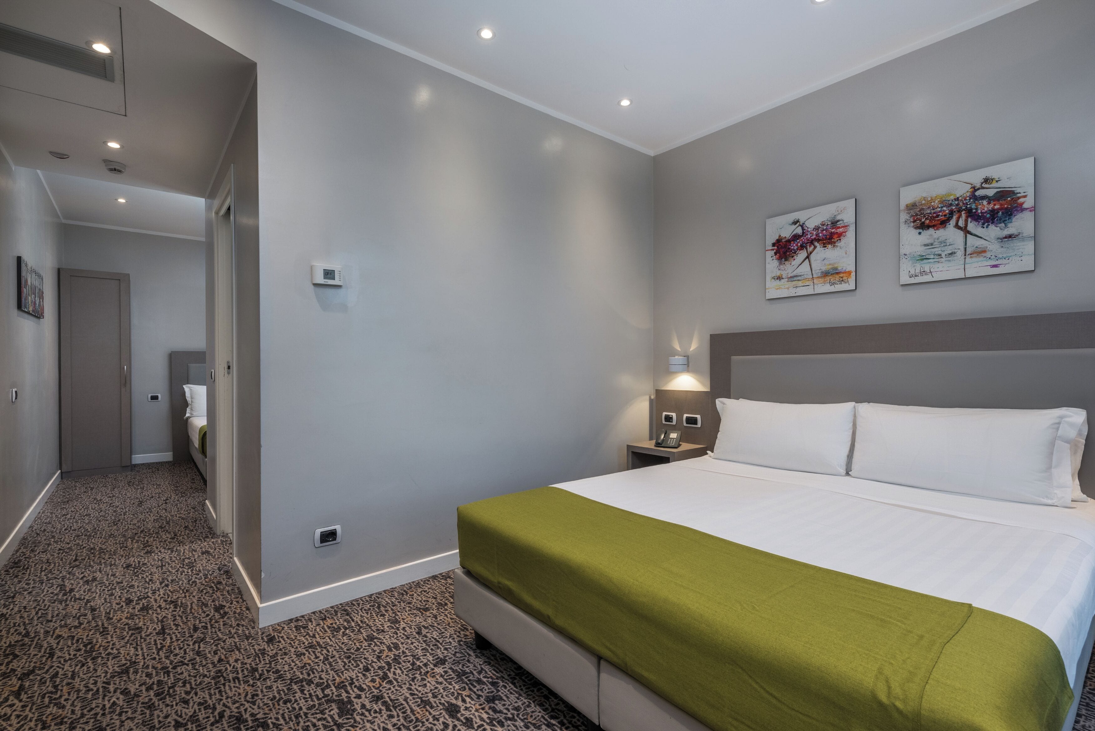 A hotel room with a large bed, a green throw, a nightstand with a lamp, and two abstract paintings on the wall.