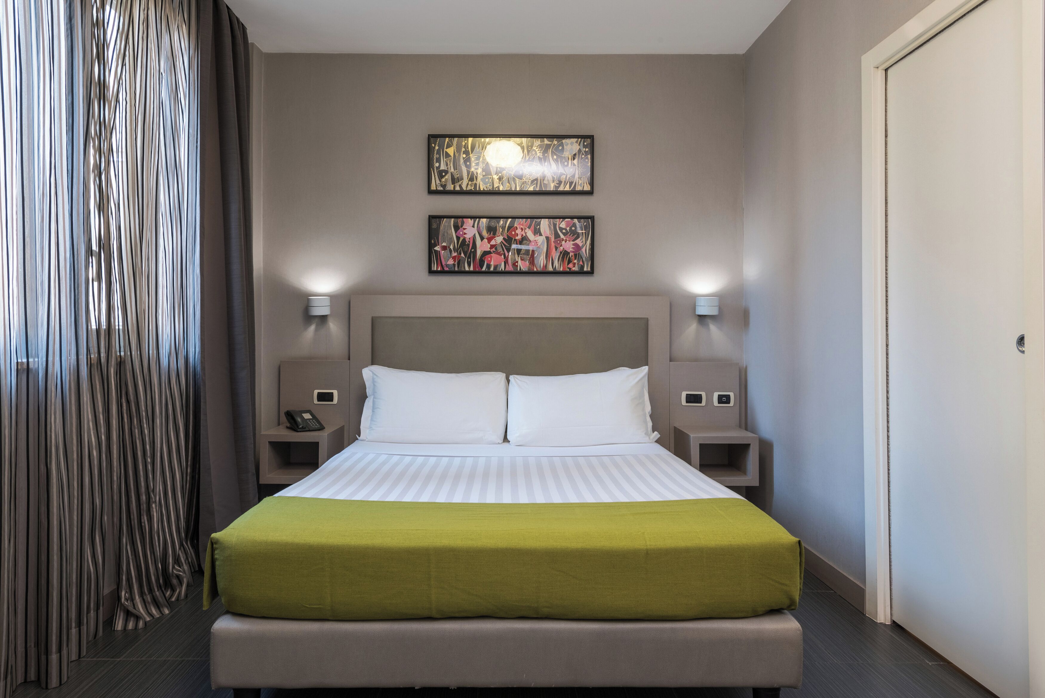 A neatly made bed with a green bedspread, two wall-mounted lamps, and two framed artworks above it.