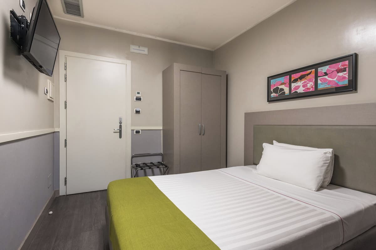 Ett hotellrum med en säng, en tv som är monterad på väggen, en garderob och en inramad tavla ovanför sängen.