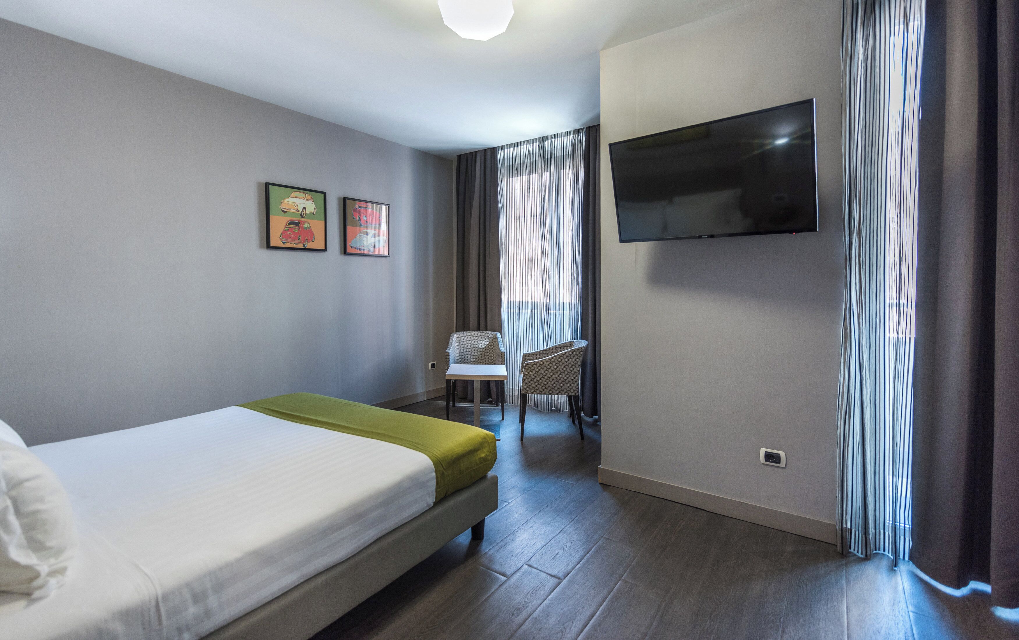 Une chambre d’hôtel avec un lit, une chaise, un téléviseur fixé au mur et des rideaux.