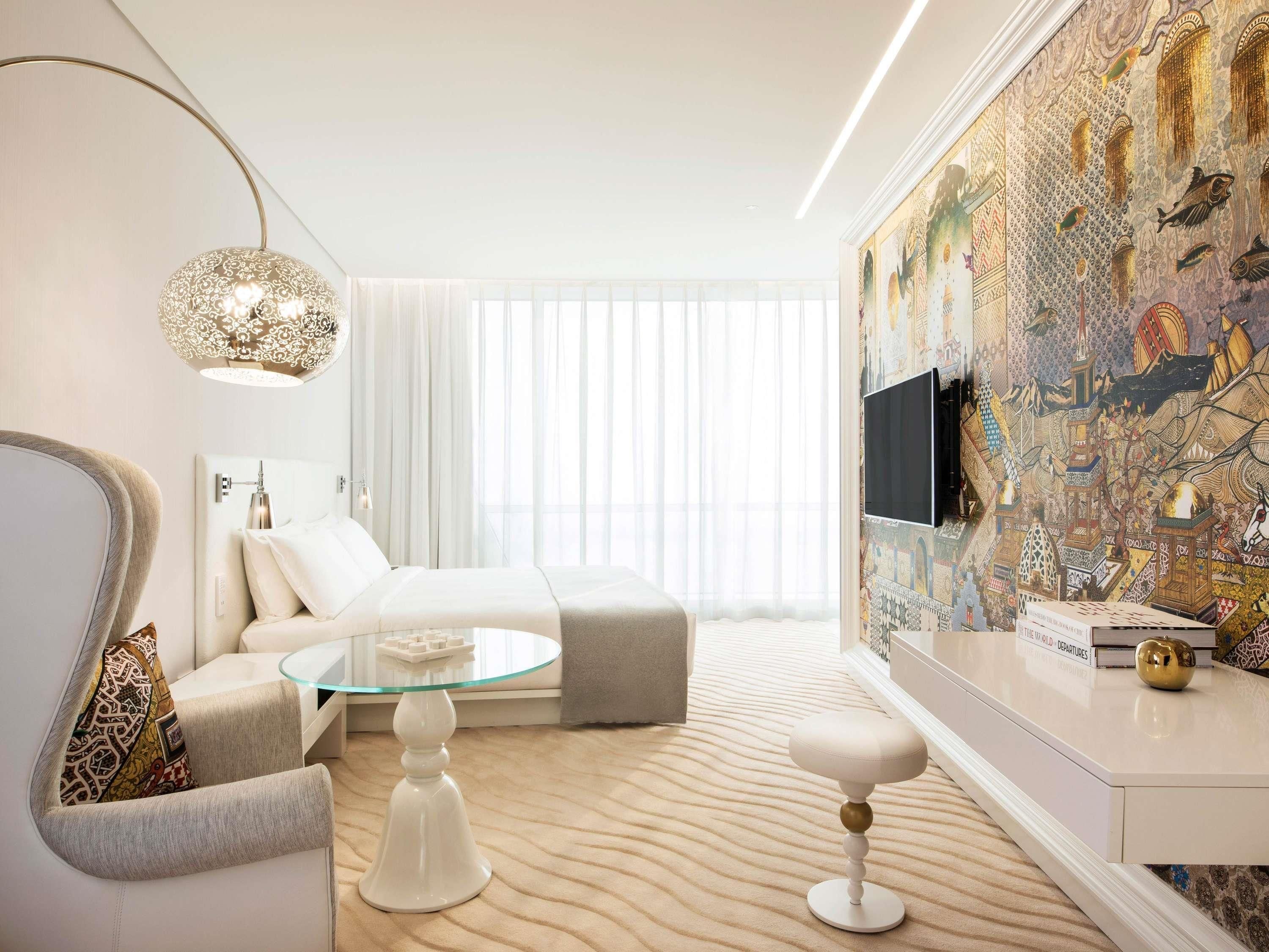 A modern hotel room with a bed, a glass coffee table, a seating area with a chair, a small table with a book, and a wall with a decorative mural.