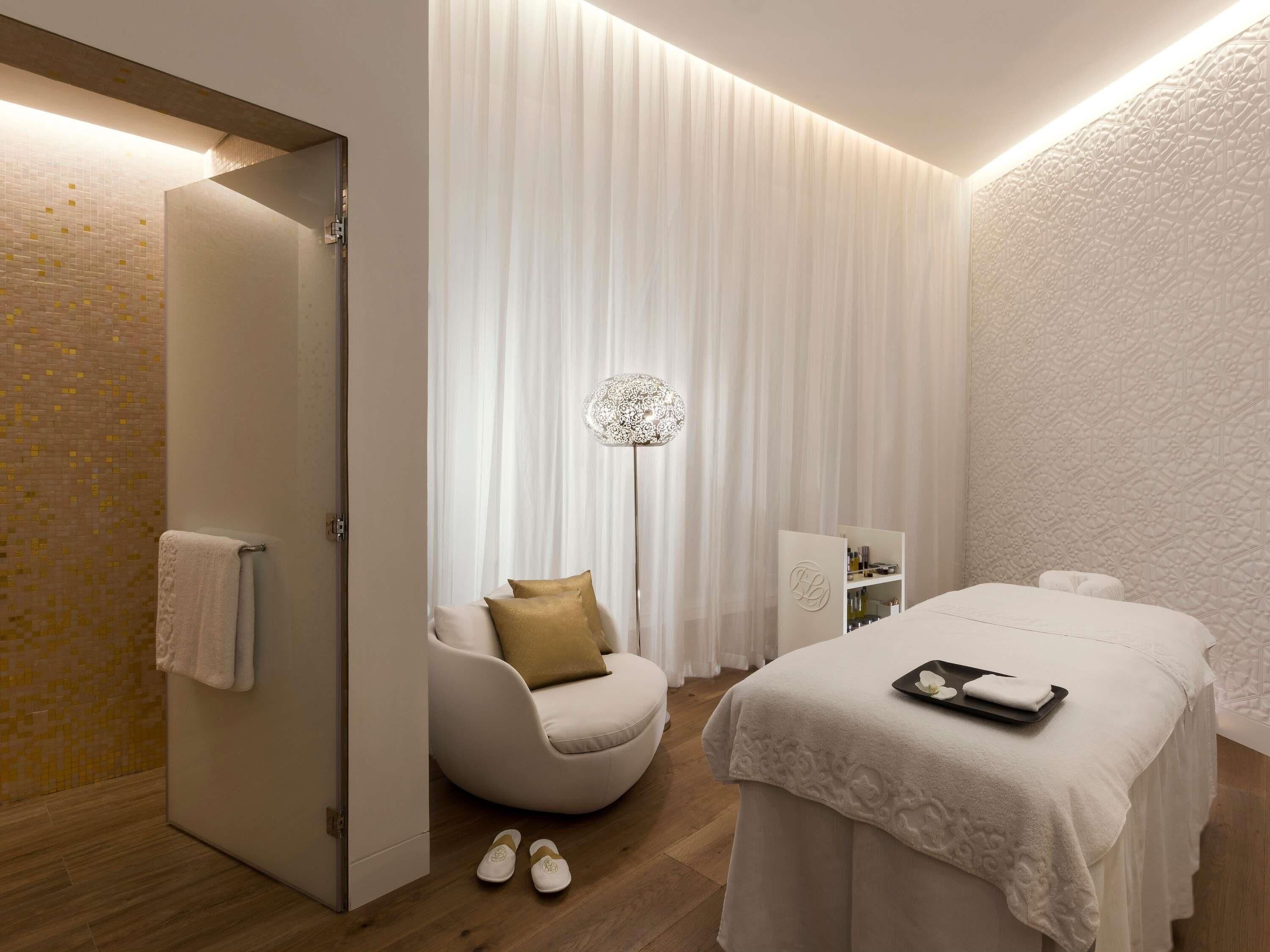 A modern spa room with a massage table, a seating area with a chair and a floor lamp, and a shower area with a towel hanging on the wall.