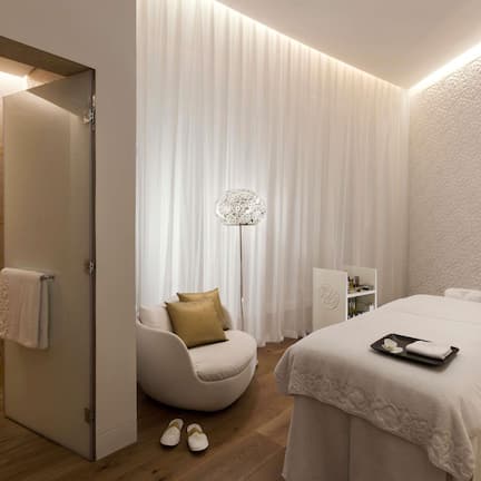 A modern spa room with a massage table, a seating area with a chair and a floor lamp, and a shower area with a towel hanging on the wall.