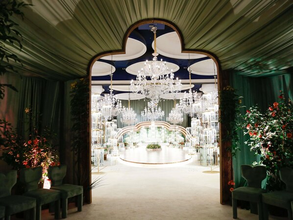 An indoor venue with a central circular stage, multiple chandeliers, and green chairs arranged on either side.