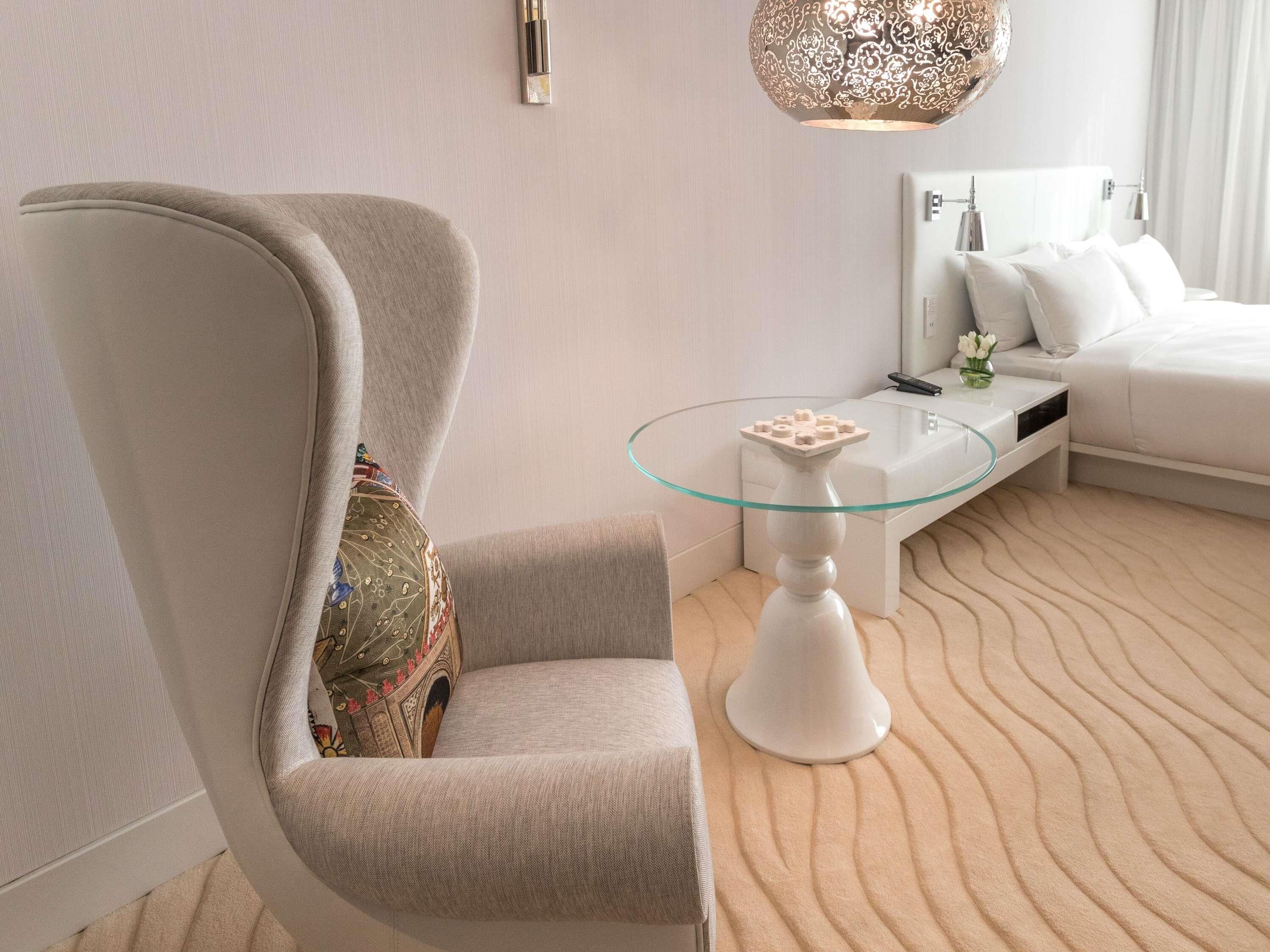 A modern hotel room with a large armchair, a glass-top table, a bed, and a decorative hanging light.