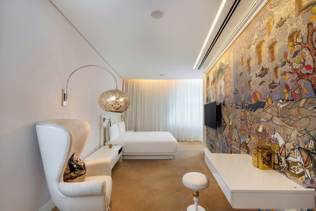 A modern bedroom with a large bed, a white armchair, a small table, and a decorative wall mural.