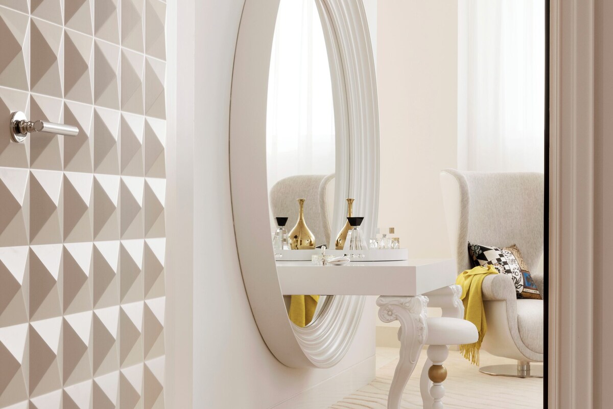 A modern bathroom with a geometric-patterned wall, a large oval mirror, and a white vanity with gold accents.