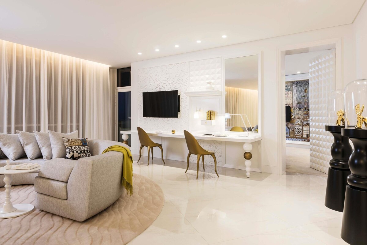 A modern living room with a sofa, a dining area with chairs, a TV mounted on the wall, and a decorative mirror.
