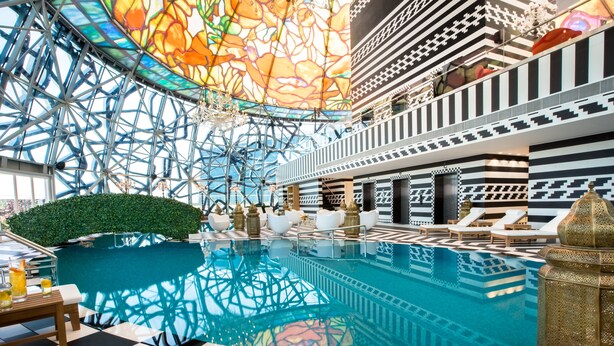 Un área de piscina interior lujosa con un diseño geométrico en el techo, suelo a cuadros y sillones de descanso.