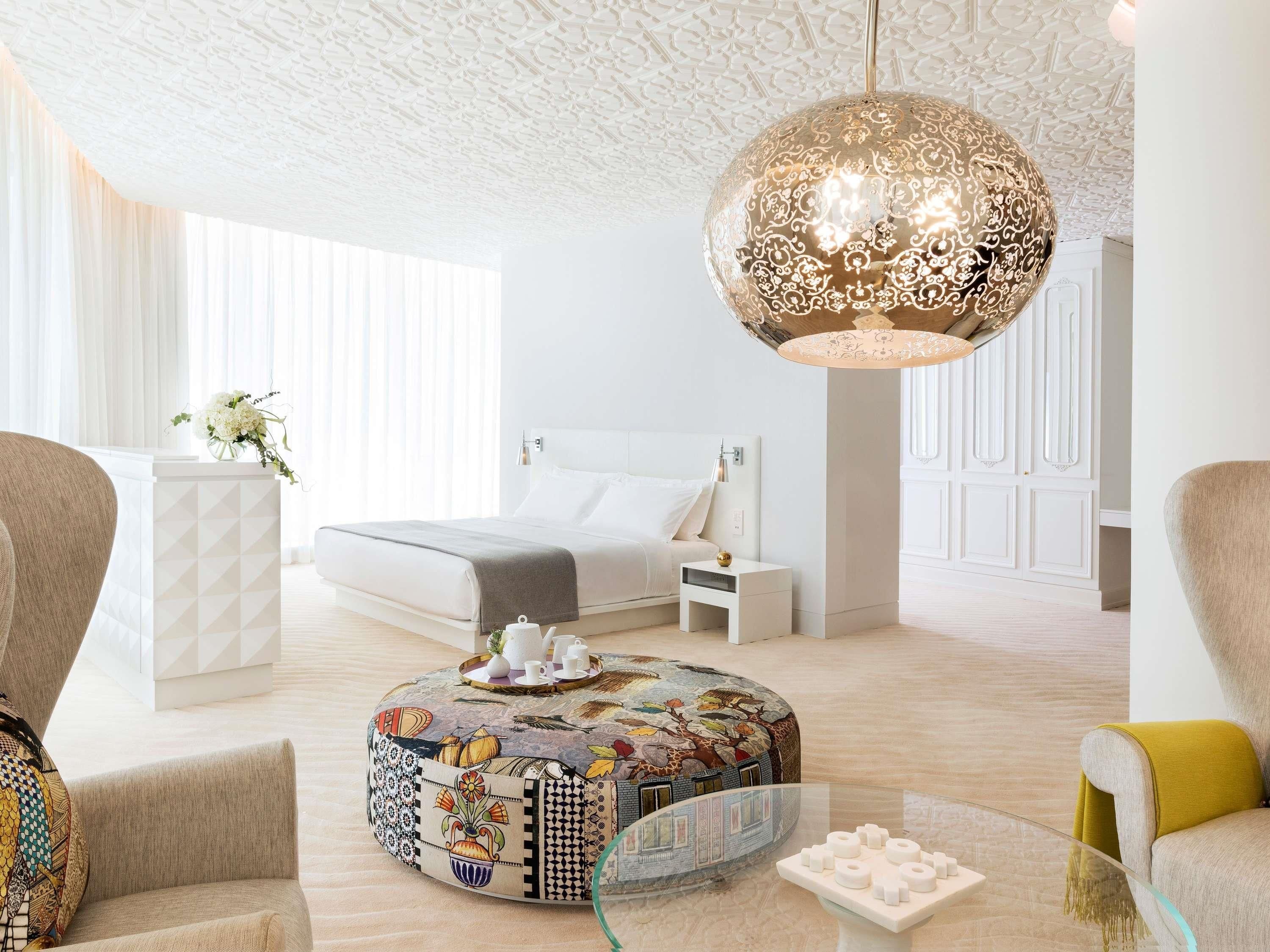 Una habitación de hotel moderna con una cama grande, un reposapiés redondo con un mosaico de colores, una mesa de centro de vidrio y una lámpara colgante.