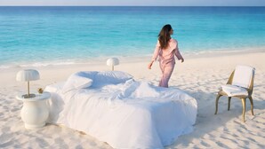Un entorno playero con una cama, dos sillas y una lámpara sobre la arena.